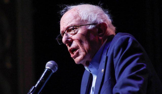 Bernie Sanders speaks passionately into a microphone on a dark stage.