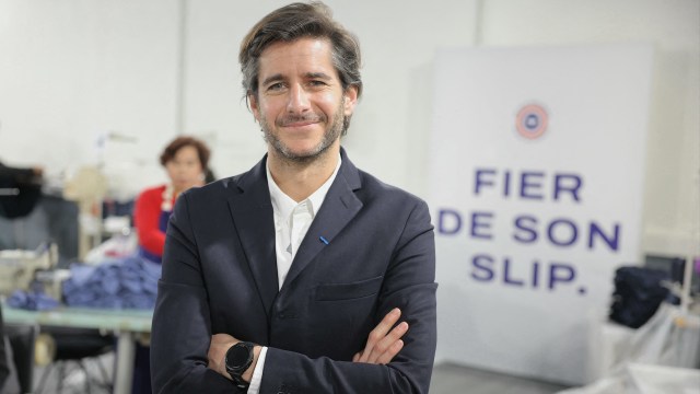 Le Slip Français CEO Guillaume Gibault smiles confidently in a workshop with a sewing machine and a sign that reads "FIER DE SON SLIP."
