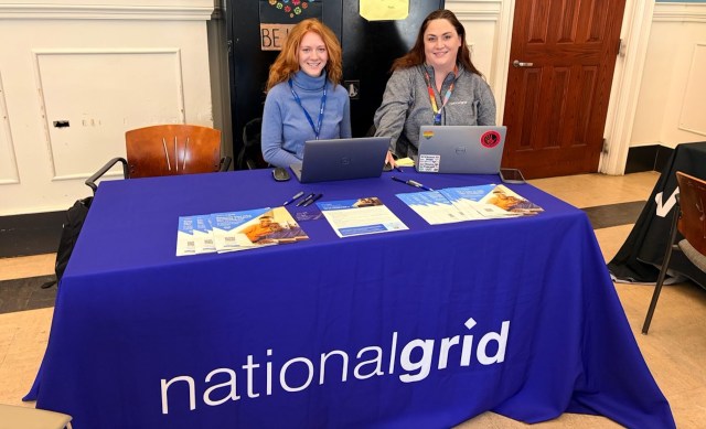 Two women seated at a table covered with a blue cloth displaying "national grid," surrounded by informational materials.