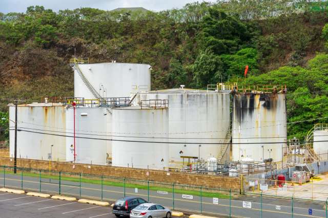 Two large white industrial storage tanks with rust and a green hillside in the background, surrounded by a fenced parking area.