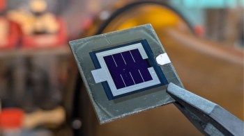 A close-up of a silicon chip with a solar cell design, held by tweezers against a blurred background.