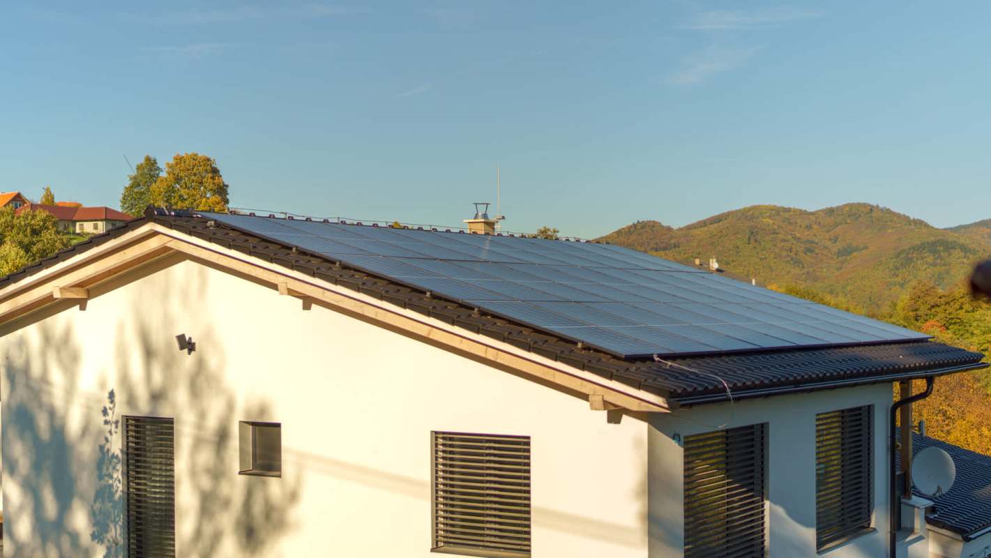 A modern house with solar panels on the roof, surrounded by green hills and a clear blue sky.
