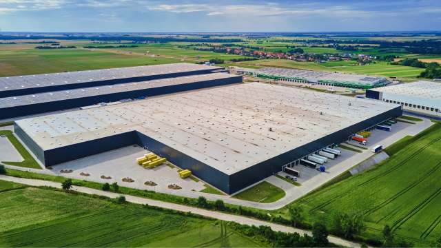 An aerial view of large industrial warehouses with green fields in the background under a partly cloudy sky.