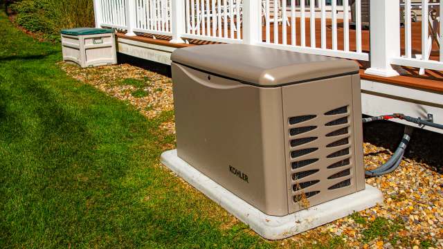 A beige generator sits on a concrete base in a grassy yard beside a wooden deck.