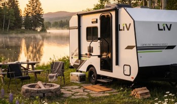 A compact camper trailer set by a serene lake, surrounded by trees and a cozy seating area with a fire pit.