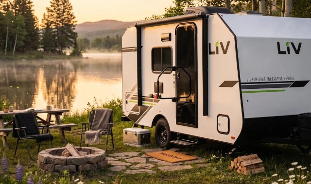 A compact camper trailer set by a serene lake, surrounded by trees and a cozy seating area with a fire pit.