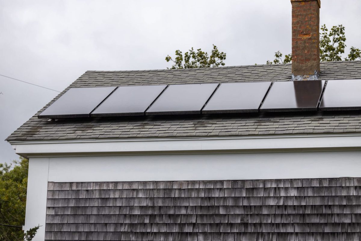 A roof with solar panels installed, featuring dark shingles and a brick chimney against a cloudy sky.