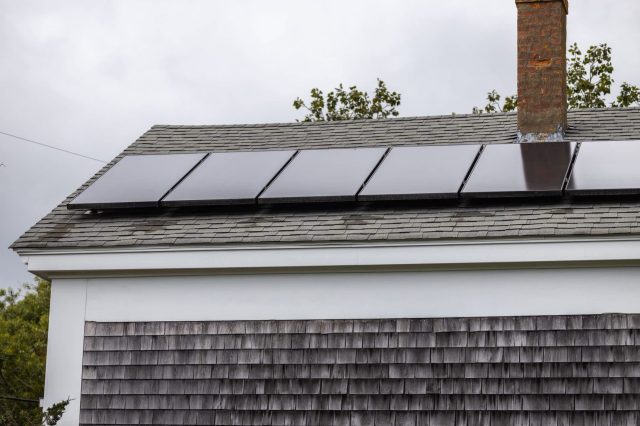 A roof with solar panels installed, featuring dark shingles and a brick chimney against a cloudy sky.