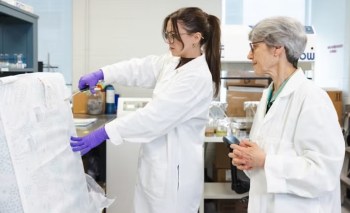 An older woman at the right in a white lab coat looks on as a younger woman cuts a sample from a mattress with scissors.