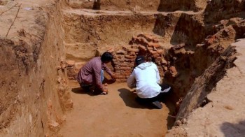 Two archaeologists examine a wall of bricks at an excavation site on a sunny day.