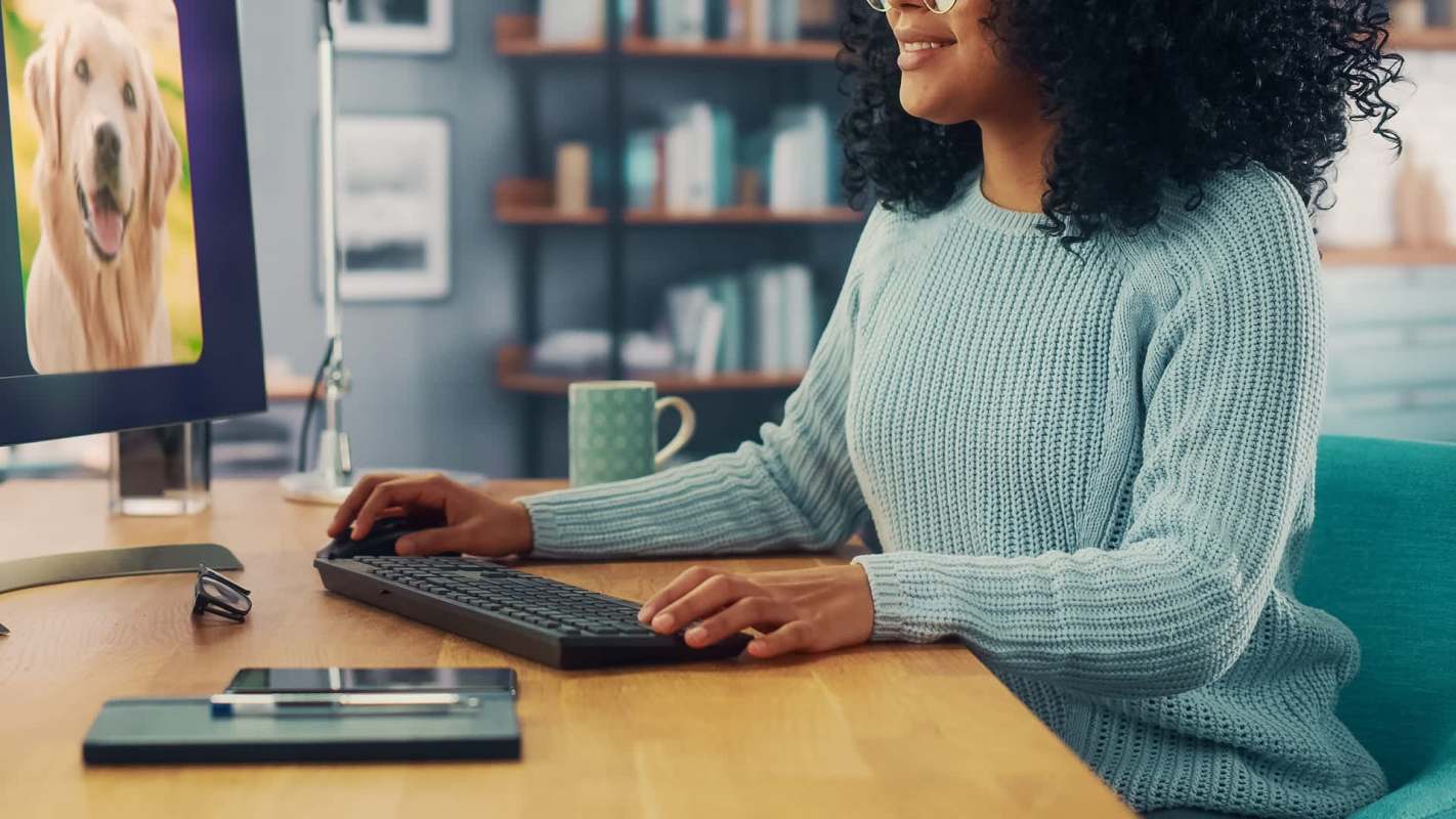 A woman with curly hair in a light sweater works at a desk with a computer displaying a dog on the screen.