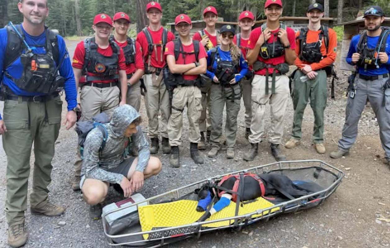 A group of individuals in rescue gear poses with a dog on a stretcher in a forested area.
