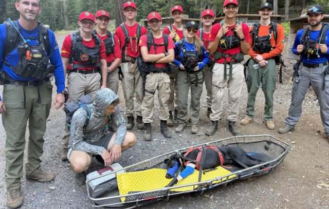 A group of individuals in rescue gear poses with a dog on a stretcher in a forested area.
