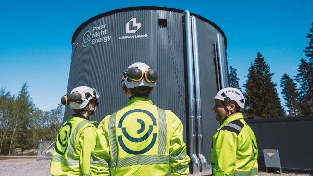 Three individuals in safety gear stand in front of a large energy storage tank with trees in the background.