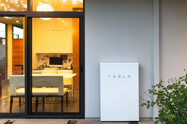 A Tesla Powerwall installed outside a modern home, with a view of a stylish dining area through glass doors.