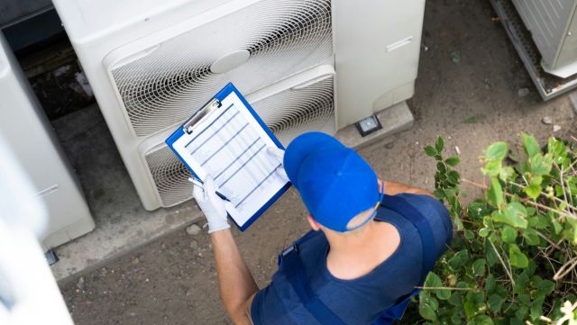 A technician wearing gloves examines a clipboard near outdoor air conditioning units.