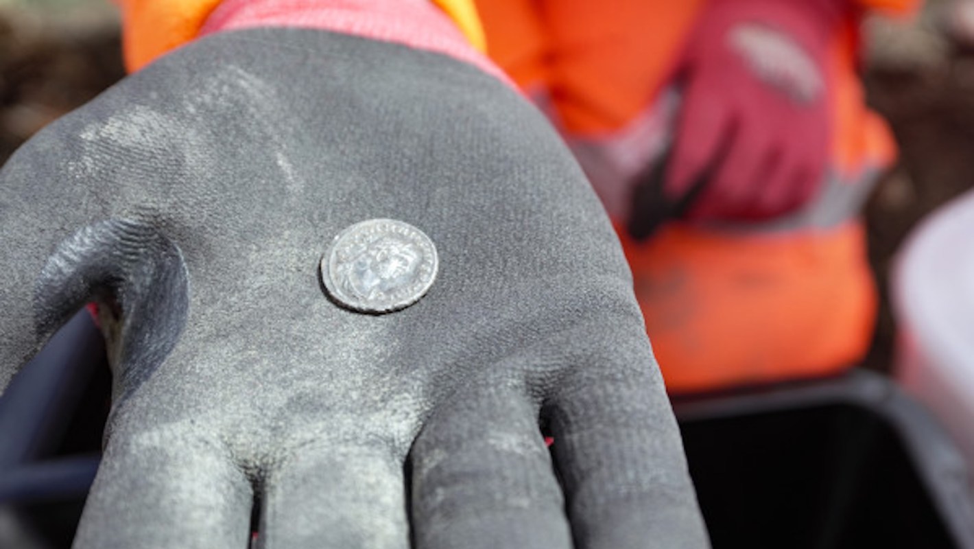 A gloved hand holds a silver coin with a detailed image, surrounded by a blurred background of workers.