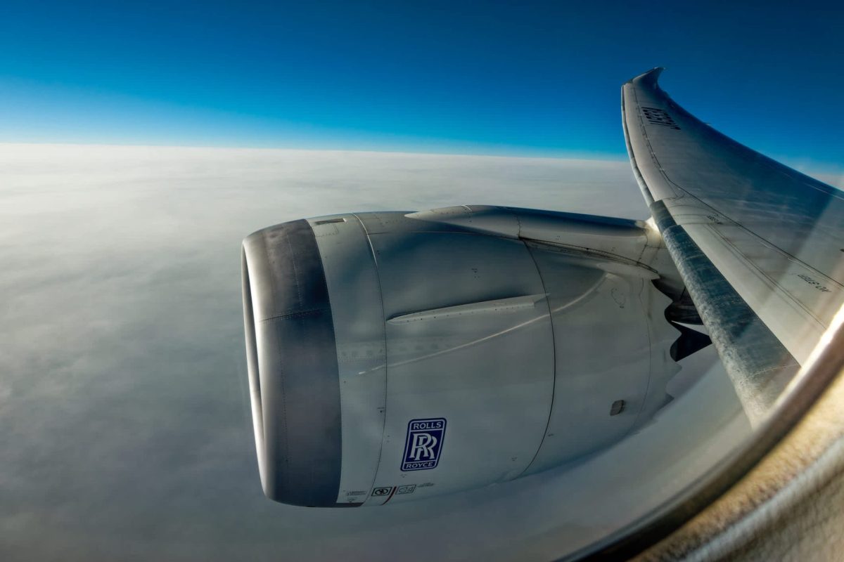 An airplane wing and engine viewed from a window, surrounded by a clear blue sky and white clouds below.