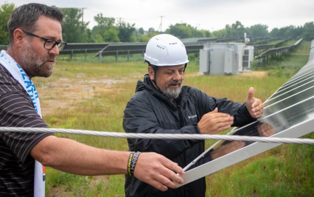 Two men, one wearing a hard hat, inspect solar panels in a field.