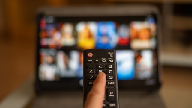 A hand holds a remote control in front of a blurred screen displaying various streaming options.