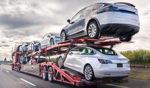A car carrier truck transporting multiple Tesla vehicles on a highway under a cloudy sky.