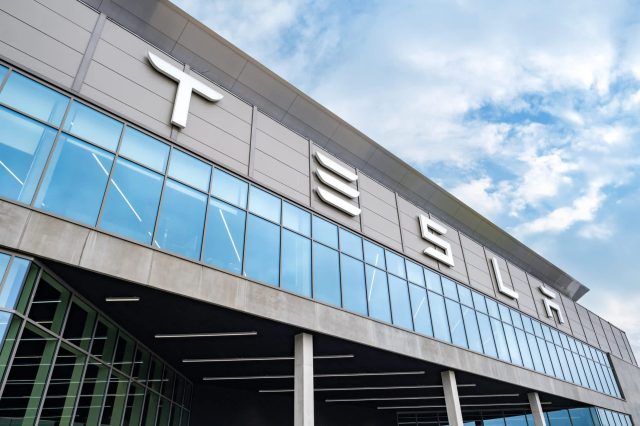 A close-up of the Tesla building exterior featuring large signage and reflective glass windows under a blue sky.