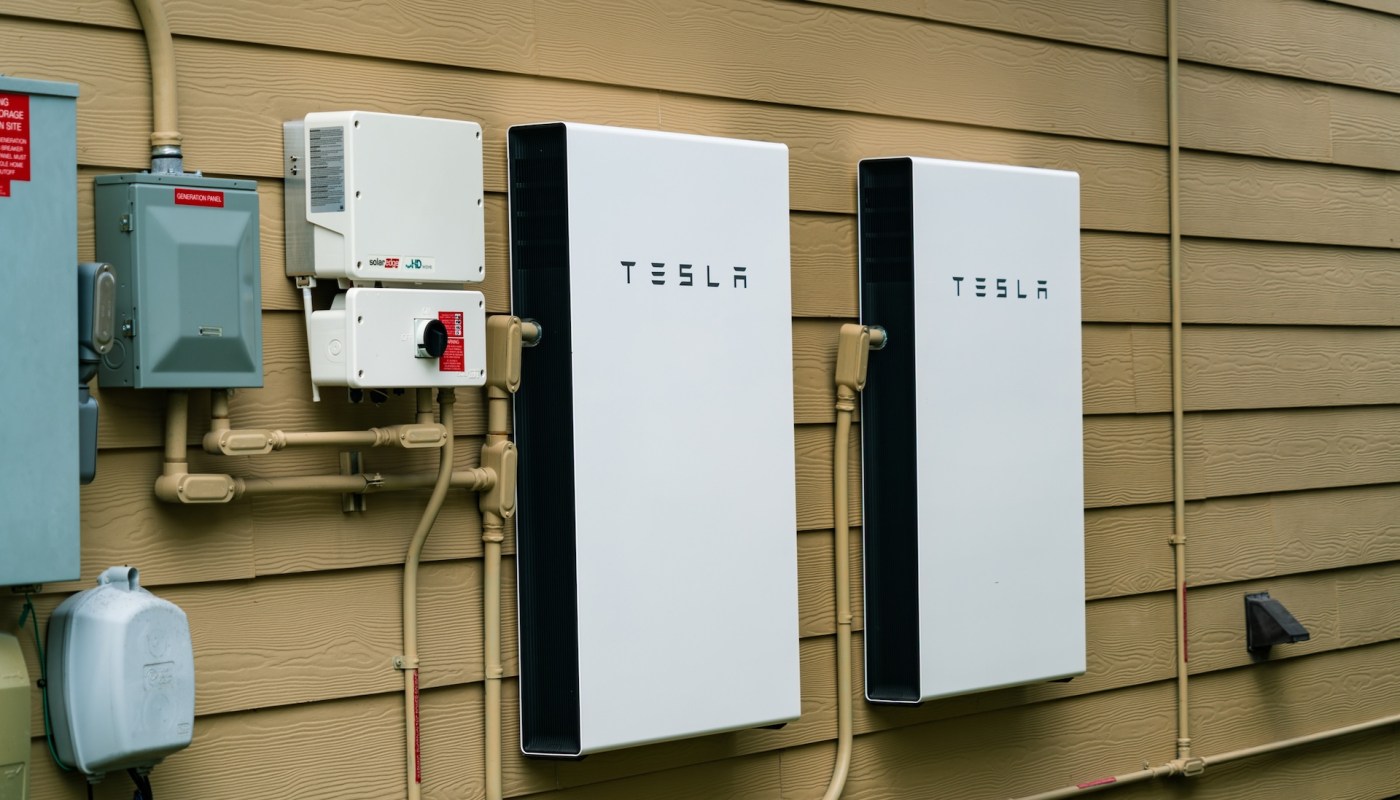 Two Tesla power wall units mounted on a wooden wall next to electrical equipment and piping.