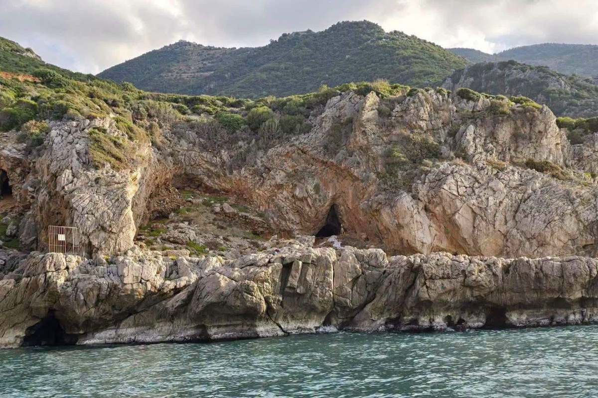 Rocky cliffs with greenery and a cave entrance, viewed from the sea under a cloudy sky.