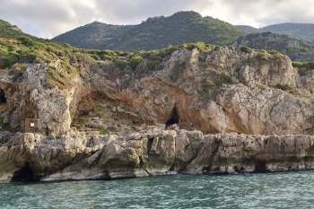 Rocky cliffs with greenery and a cave entrance, viewed from the sea under a cloudy sky.
