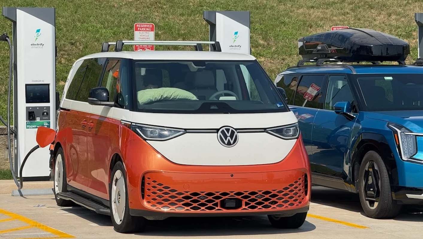 A Volkswagen electric vehicle with an orange and white design is charging at an electric station alongside another car.