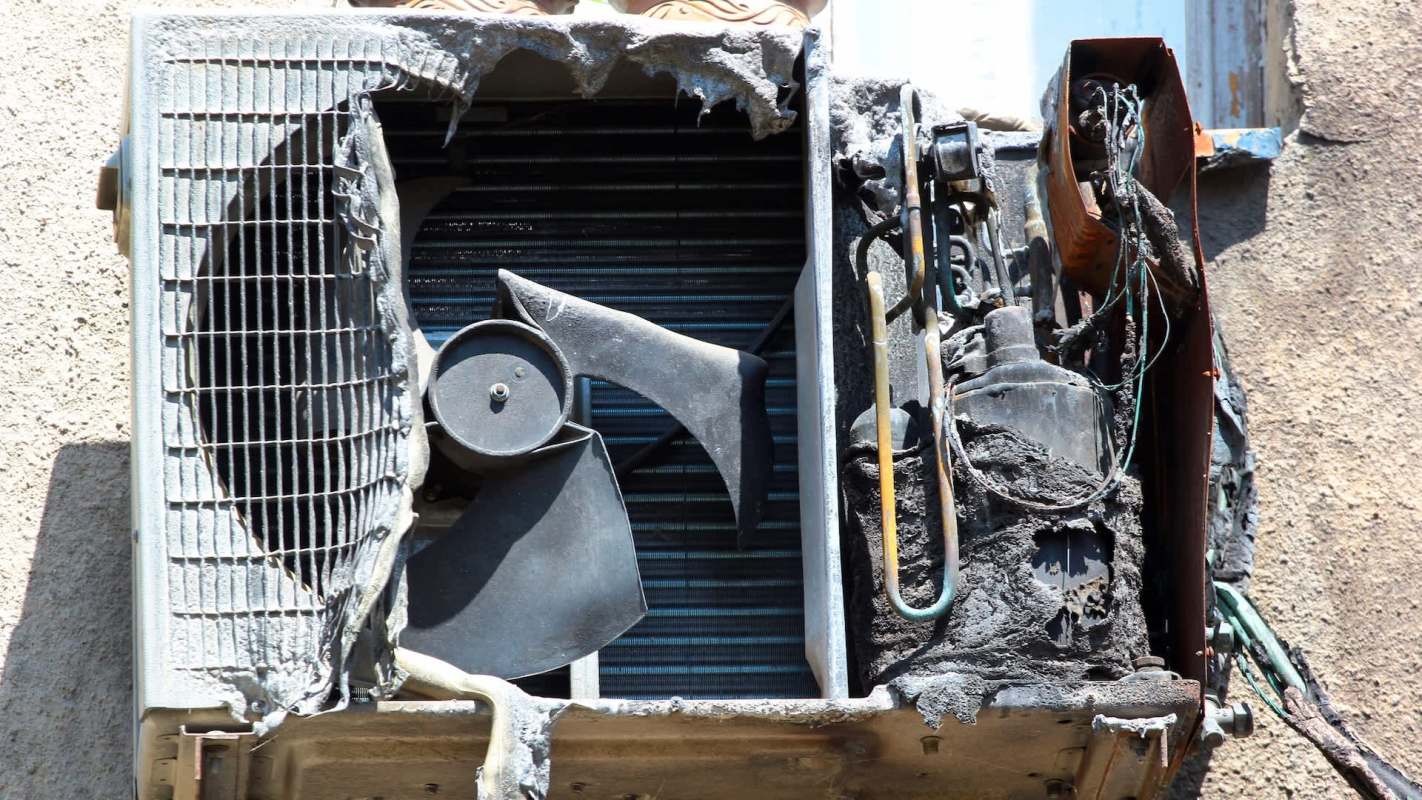 A damaged air conditioning unit with charred components, wires exposed, and a missing protective cover.
