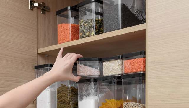A hand reaching for a transparent container filled with pink salt in a well-organized kitchen cabinet.