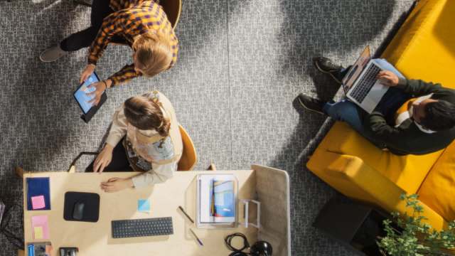 Three people collaborate in a bright, modern workspace, using a tablet and laptop while seated at a wooden table.