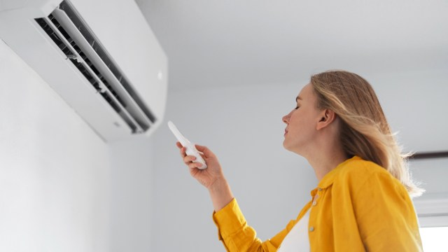 A woman in a yellow shirt uses a remote control to adjust a wall-mounted air conditioner.