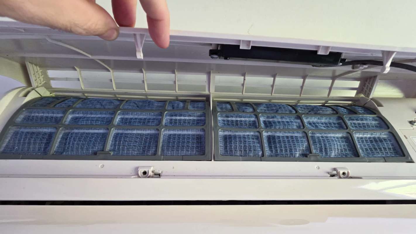 A person holds open the cover of an air conditioning unit, revealing its blue mesh filters.