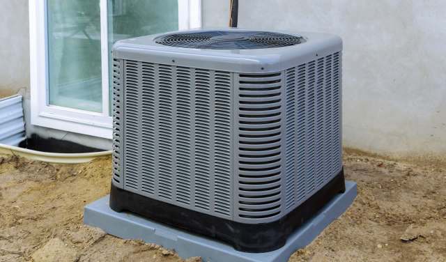 An outdoor air conditioning unit is positioned beside a window, set on a concrete base against a dirt background.