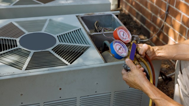 A technician holds gauges and hoses while servicing an outdoor air conditioning unit.