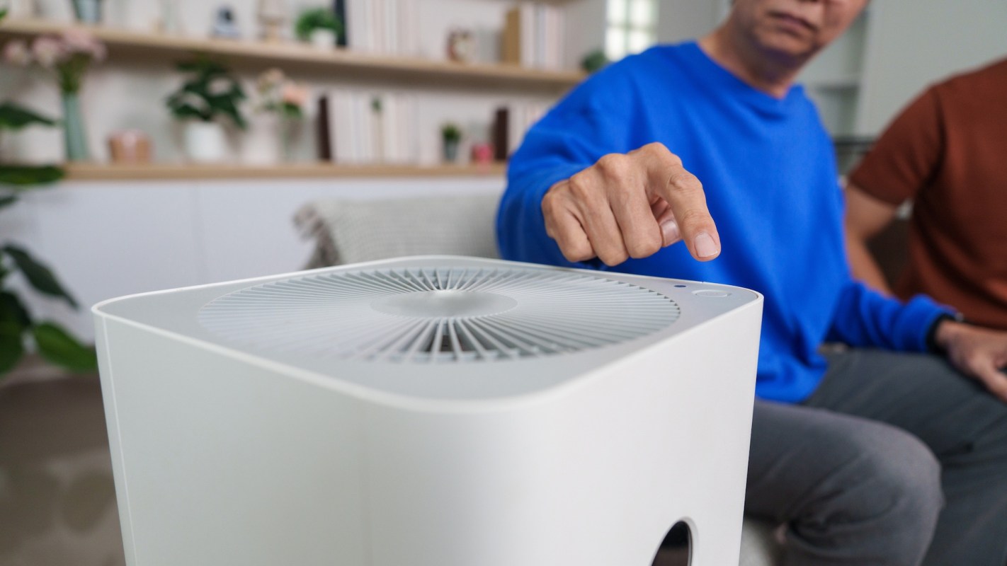 A person points at a white air purifier in a spacious, green indoor setting.