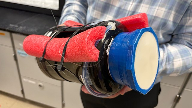 A person holds a black cylindrical device with red foam and blue plastic components in a laboratory setting.