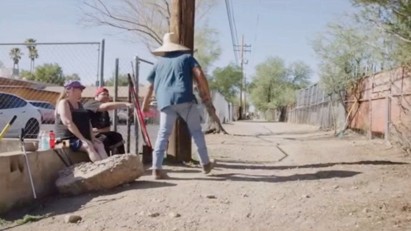 A person in a wide-brimmed hat walks down a dirt alley.