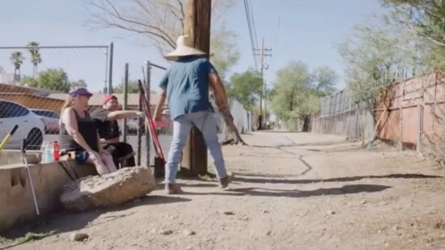 A person in a wide-brimmed hat walks down a dirt alley.