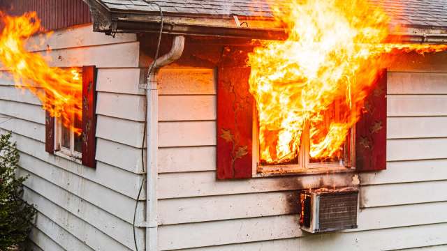 Flames engulf a window of a small house, with smoke billowing from an air conditioning unit below.