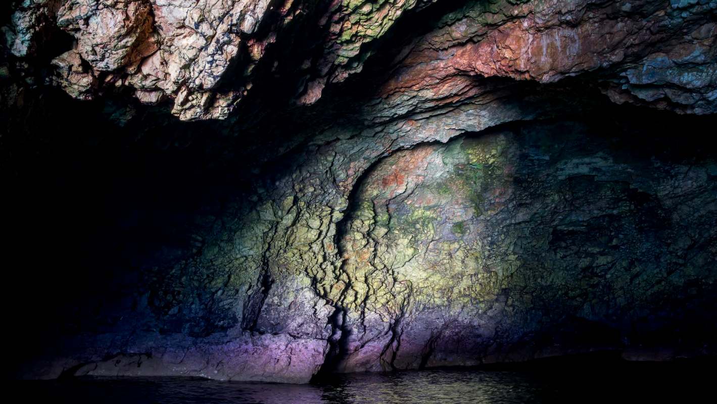 An illuminated cave with colorful walls rising from the water.