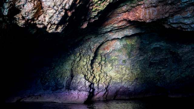 An illuminated cave with colorful walls rising from the water.