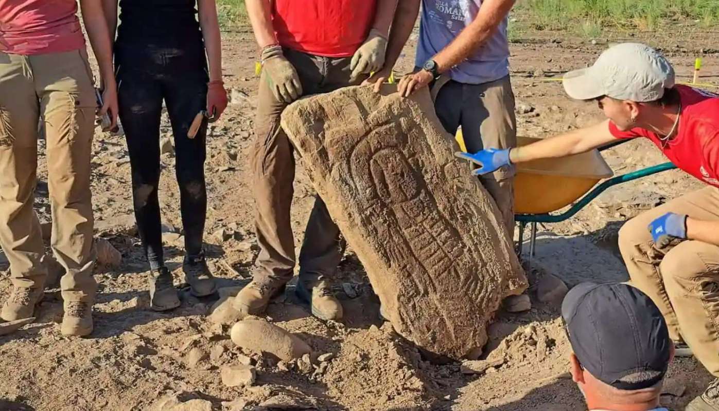 A group of archaeologists carefully handle a large carved stone while working at a dig site.
