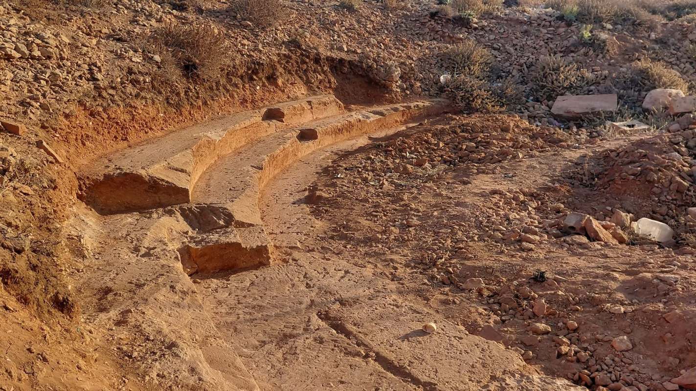A curved excavation in reddish soil, featuring large stone blocks and sparse vegetation nearby.