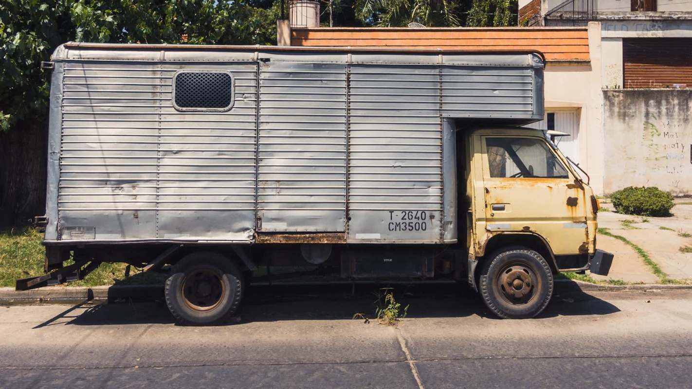 An old, rusted truck with a metallic cargo area parked on the street near a building.