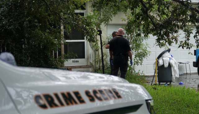 Two police officers investigate outside a house with a trash can nearby marked with a crime scene vehicle in the foreground.