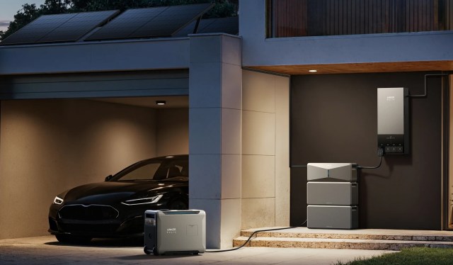 A sleek black electric vehicle parked in a garage with solar panels, a power station, and battery storage setup.