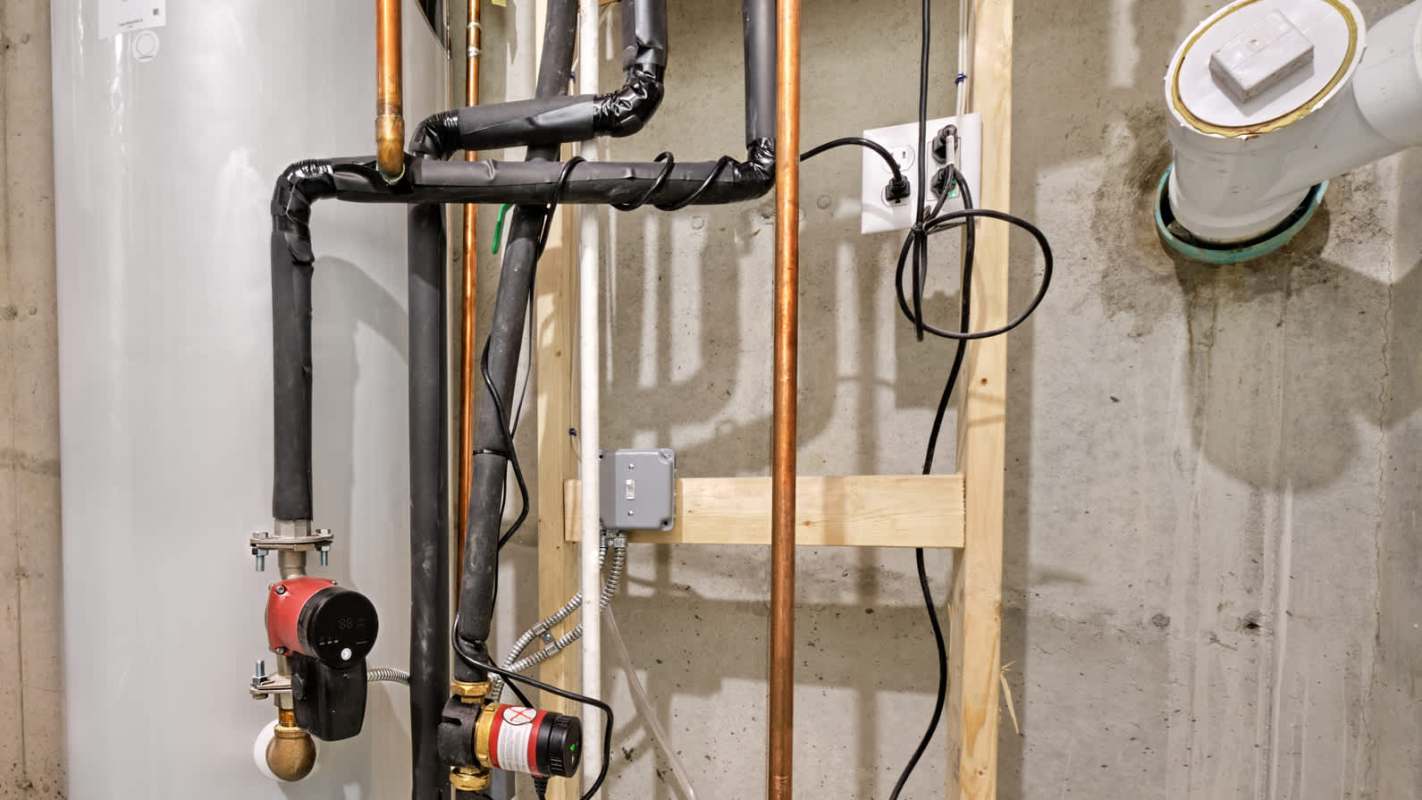 Pipes and valves connected to a water heater in a basement, with electrical components mounted on a wooden frame.